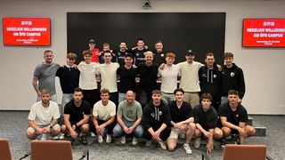 Eine Gruppe von Männern in Sportkleidung posiert für ein Foto in einem Raum mit einer Tafel, einige sitzend und andere stehend.