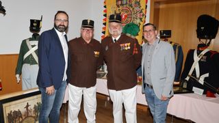 Vier Männer in einem Raum, zwei in Militäruniformen und zwei in Freizeitkleidung, posieren für ein Foto. Ein Flagge und ein Tisch mit Objekten im Hintergrund.