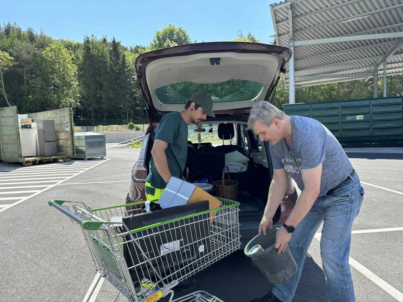 Zwei Männer entladen Gegenstände aus dem Kofferraum eines Autos, einer hält einen Eimer und der andere einen Einkaufswagen. Sie sind von Gegenständen wie Kisten, einem Korb und einer Aufbewahrungsbox umgeben.