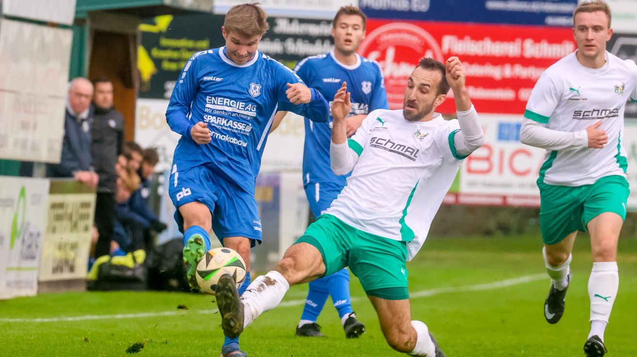 Fußballspieler in blauen und grünen Trikots auf einem Feld, einer tritt den Ball, während der andere zusieht. Hinter ihnen steht ein Spieler in Blau.