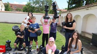 Eine Gruppe von Kindern und Erwachsenen posiert mit riesigen Schachfiguren in einem Innenhof. Einige tragen Freizeitkleidung, andere T-Shirts. Die Umgebung hat ein Gebäude mit Bogenfenstern und einem Rasen.