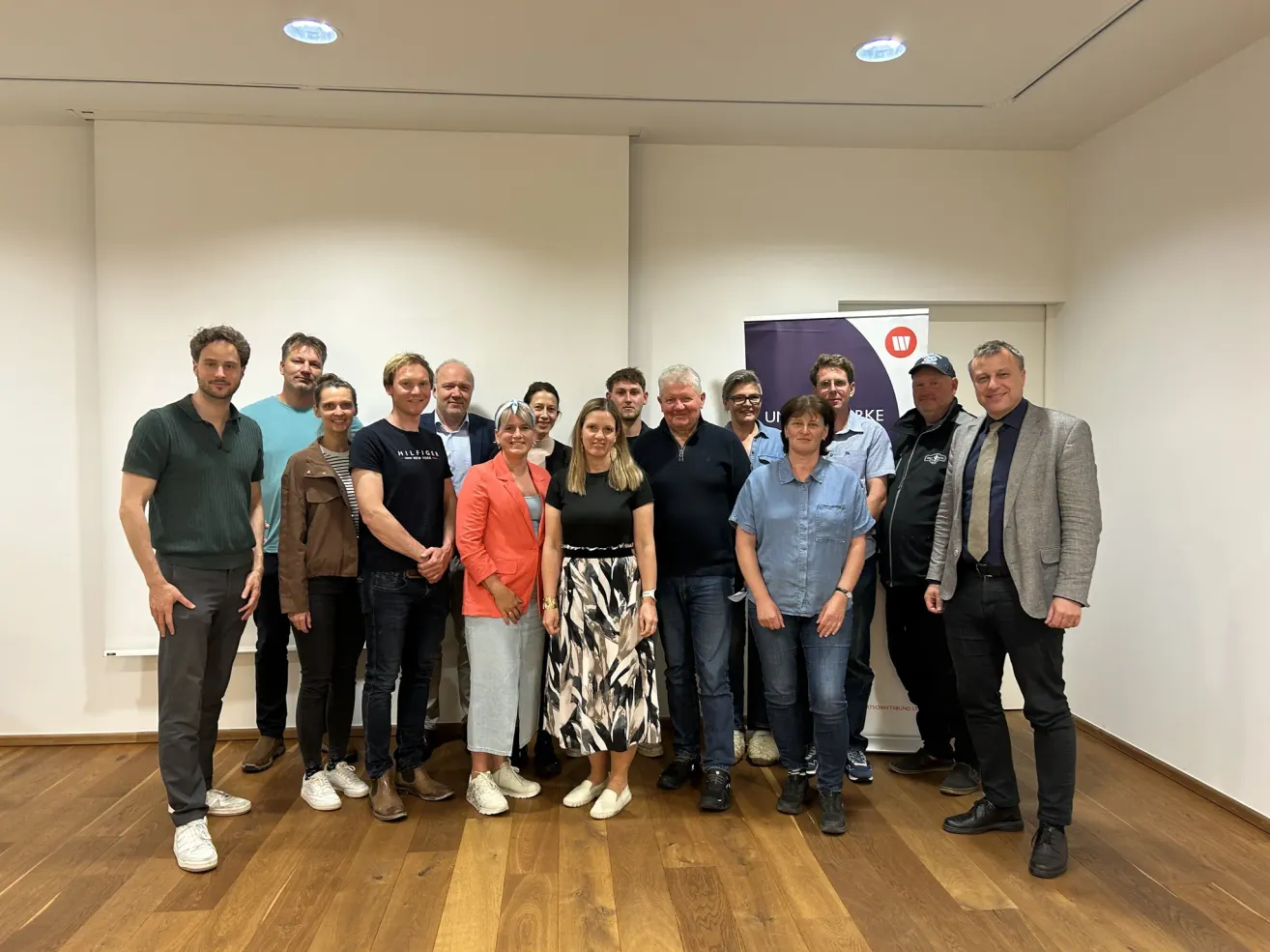 Eine Gruppe von Menschen steht für ein Foto zusammen und lächelt, in einem Raum mit weißen Wänden, Holzboden und einem Banner.