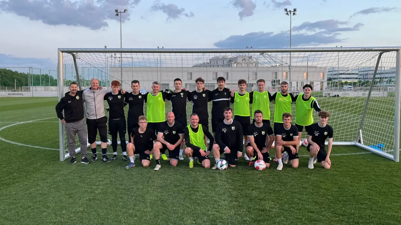 Eine Fußballmannschaft posiert für ein Gruppenfoto vor einem Netz, wobei einige Spieler grüne Westen und andere schwarze tragen. Sie lächeln und posieren mit Fußballbällen auf dem Feld.