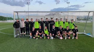 Eine Fußballmannschaft posiert für ein Gruppenfoto vor einem Netz, wobei einige Spieler grüne Westen und andere schwarze tragen. Sie lächeln und posieren mit Fußballbällen auf dem Feld.