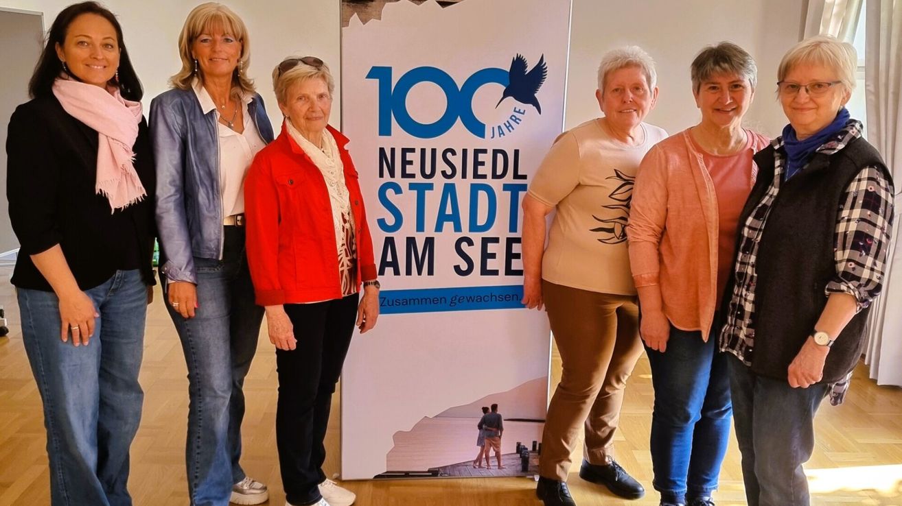 Fünf Frauen posieren vor einem Banner, das 100 Jahre Neusiedel Stadt Am See sagt. Sie alle lächeln und schauen in die Kamera.