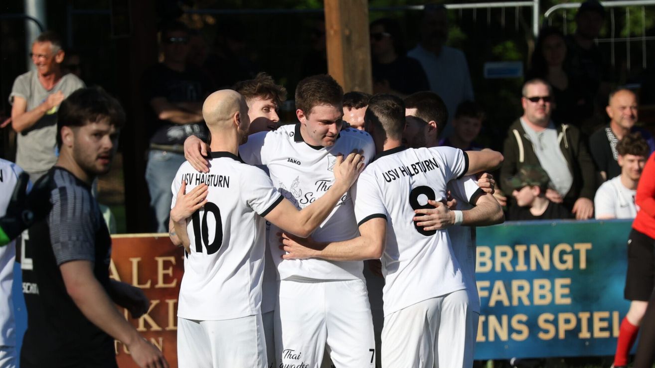 Fußballspieler in weißen Trikots mit den Nummern 10, 7 und 8 umarmen sich gegenseitig, mit einer Menge im Hintergrund.