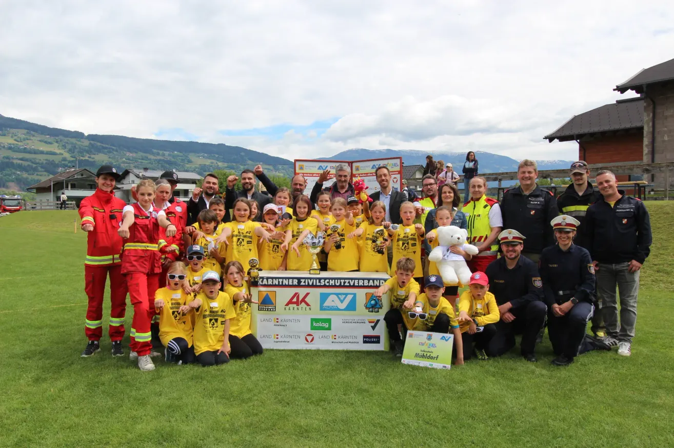 Eine Gruppe von Menschen in gelben Uniformen posiert für ein Foto mit einem großen Banner. Sie stehen auf einem grünen Feld mit Bergen im Hintergrund.