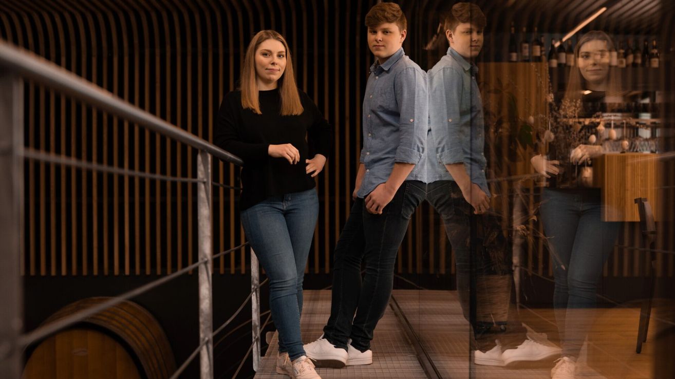 Eine Frau und zwei Männer stehen gemeinsam auf einer Plattform und posieren für ein Foto. Die Frau trägt ein schwarzes Shirt und Jeans. Die Männer tragen hellblaue Hemden und dunkle Hosen.