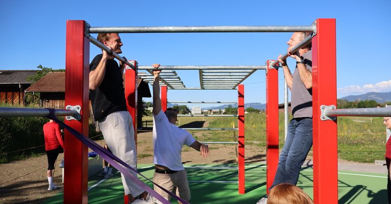 Three men are doing pull-ups on a red and gray bar in an outdoor area with a clear sky.