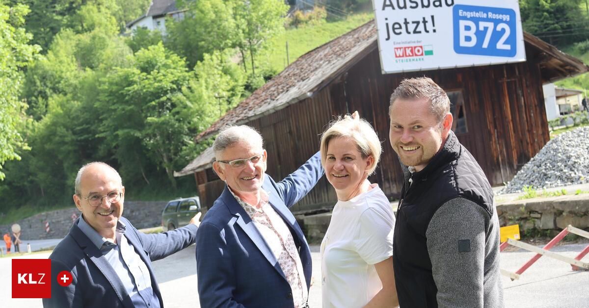 Vier Personen stehen vor einem rustikalen Holzhaus. Sie lächeln und scheinen für ein Foto zu posieren. Das Haus hat ein Schild mit der Aufschrift 'Ausbau jetzt!' mit 'B' und 'WKO' darauf.