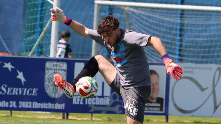 Ein Fußballspieler in einem grauen Trikot und Shorts tritt einen bunten Ball während eines Spiels. Er trägt Handschuhe und Turnschuhe. Ein unscharfes Bild eines anderen Spielers und eines Banners im Hintergrund.