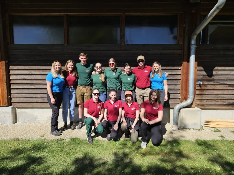 Eine Gruppe von Menschen in grünen und roten Shirts posiert für ein Foto vor einem Holzgebäude. Das Wetter ist sonnig.