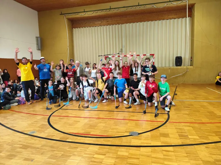 Eine Gruppe von Kindern und Erwachsenen posiert mit Hockeyschlägern auf einem Basketballplatz für ein Foto.