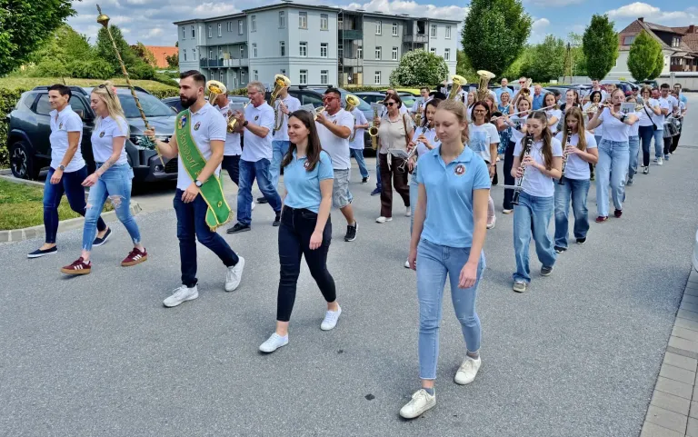 Eine Blaskapelle aus Männern und Frauen, in blauen Uniformen, zieht mit Instrumenten in einer Parade. Sie werden von Autos und Gebäuden im Hintergrund begleitet.