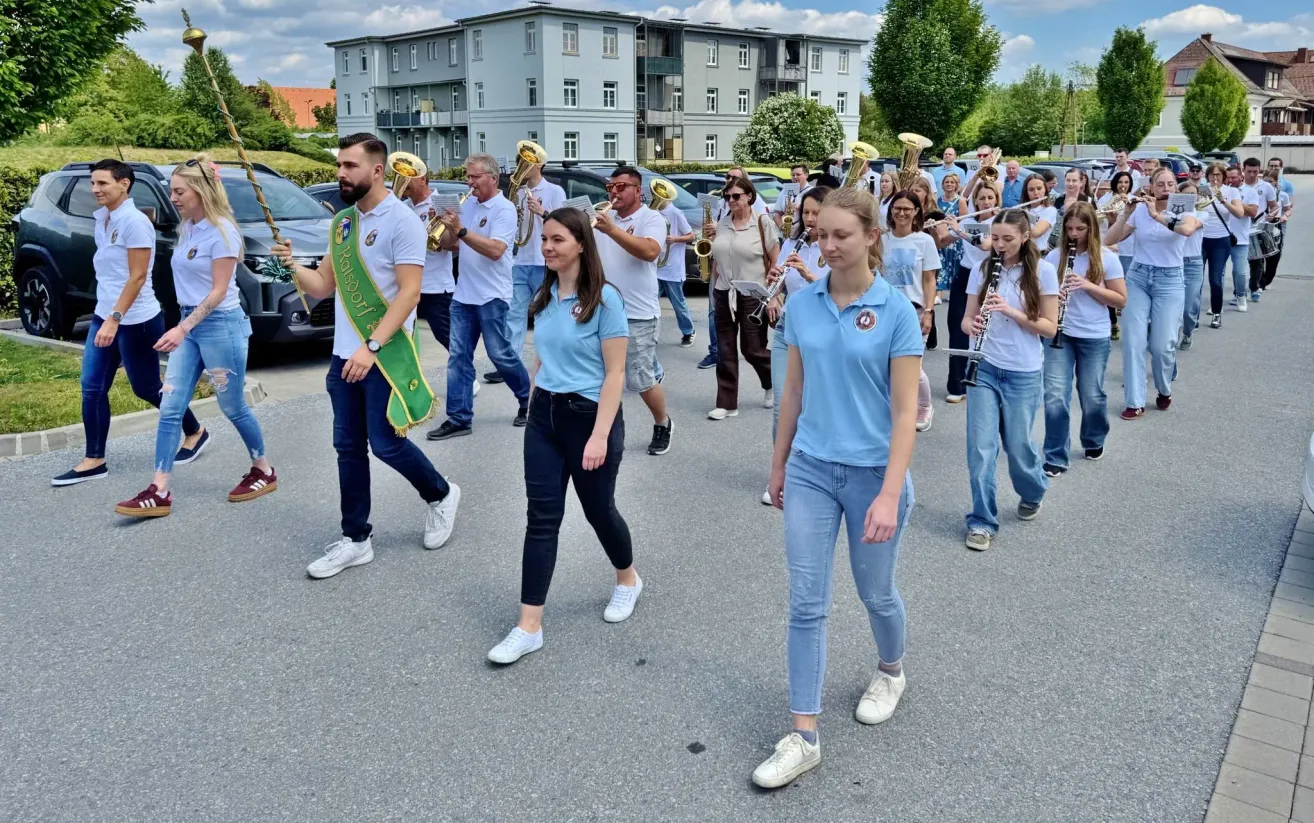 Eine Blaskapelle aus Männern und Frauen, in blauen Uniformen, zieht mit Instrumenten in einer Parade. Sie werden von Autos und Gebäuden im Hintergrund begleitet.