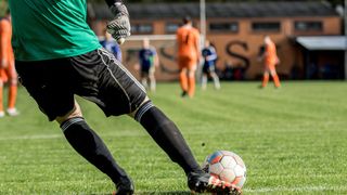 Ein Fußballspieler in grüner und schwarzer Ausrüstung ist dabei, einen Fußball zu schießen. Andere Spieler und ein Gebäude sind im Hintergrund zu sehen.