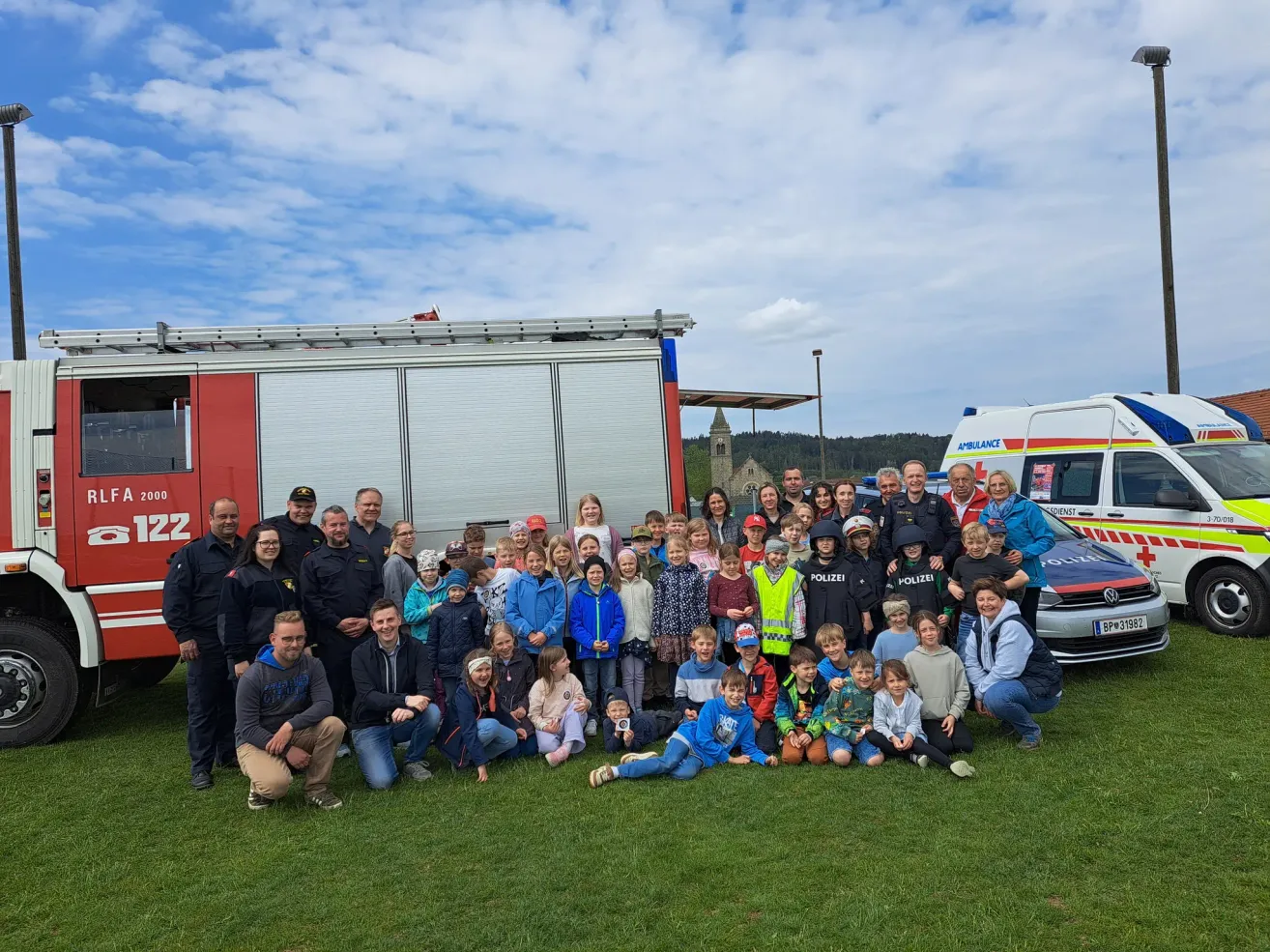 Gruppenfoto mit Feuerwehrleuten, Polizisten und Kindern an einem sonnigen Tag vor Rettungsfahrzeugen.