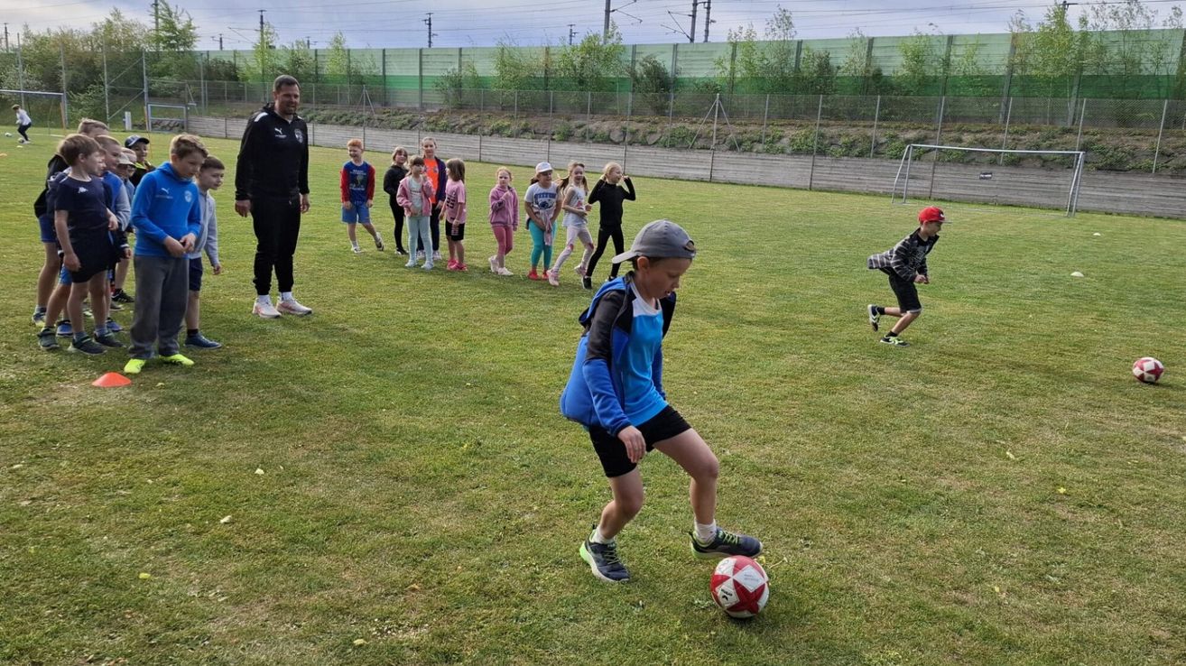 Ein Junge tritt einen Fußball auf einem grünen Feld. Kinder stellen sich hinter einem Zaun auf, und ein erwachsener Mann steht in der Nähe.