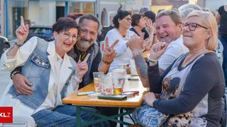 Eine Gruppe von Menschen sitzt an einem Tisch im Freien mit Getränken und einem Handy. Sie lächeln und geben ein Friedenszeichen. Dahinter sind verschwommene Menschen und Gebäude.
