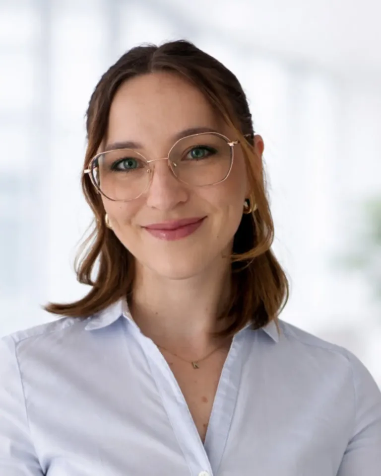 Eine Frau mit braunen Haaren und grünen Augen, die eine Brille und ein weißes Hemd trägt, lächelt in die Kamera.