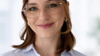 Eine Frau mit braunen Haaren und grünen Augen, die eine Brille und ein weißes Hemd trägt, lächelt in die Kamera.