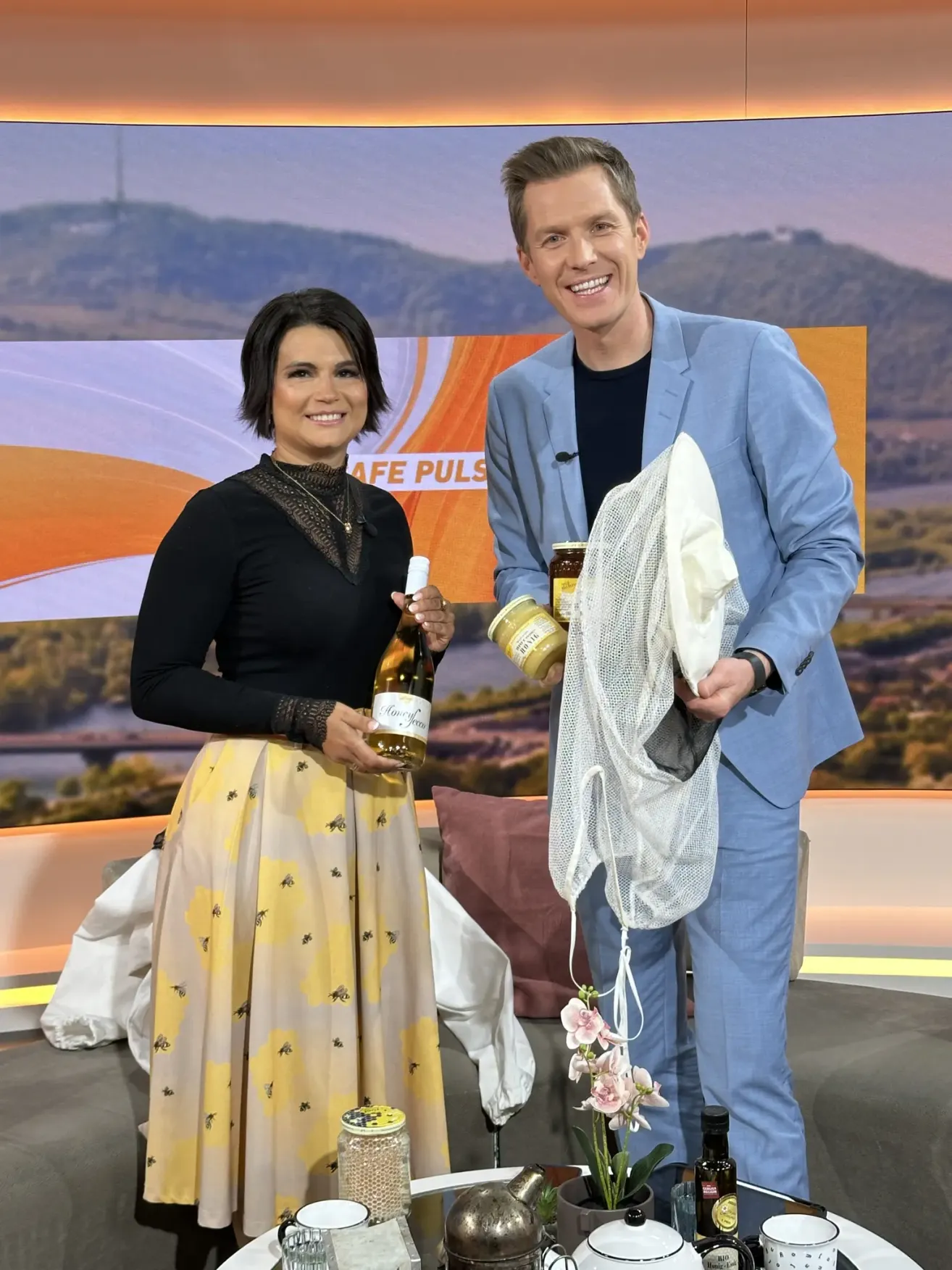 Eine Frau in einem Blumenrock und ein Mann in einem blauen Anzug stehen lächelnd auf einer Bühne mit einem Hintergrund von Bergen. Die Frau hält eine Weinflasche und der Mann hält einen Topf und ein Netz.