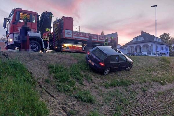 Ein dunkles Auto ist auf einem grasbewachsenen Hügel beschädigt, mit einem Feuerwehrwagen in der Nähe. Feuerwehrleute beurteilen den Schaden.