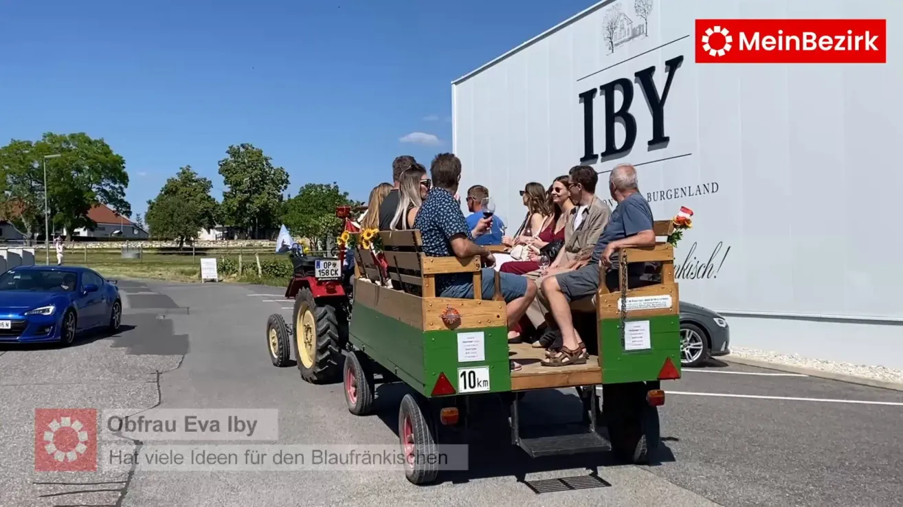 Eine Gruppe von Menschen fährt in einem von einem Traktor gezogenen Wagen, mit einem Gebäude, das mit IBY beschriftet ist, im Hintergrund. Sie halten Weingläser und genießen die Fahrt.