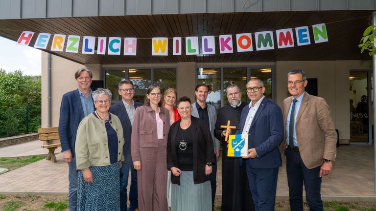 Eine Gruppe von Menschen steht vor einem Gebäude mit einem Banner, auf dem 'LICH WILLKOMM' steht. Sie lächeln und posieren für ein Foto.