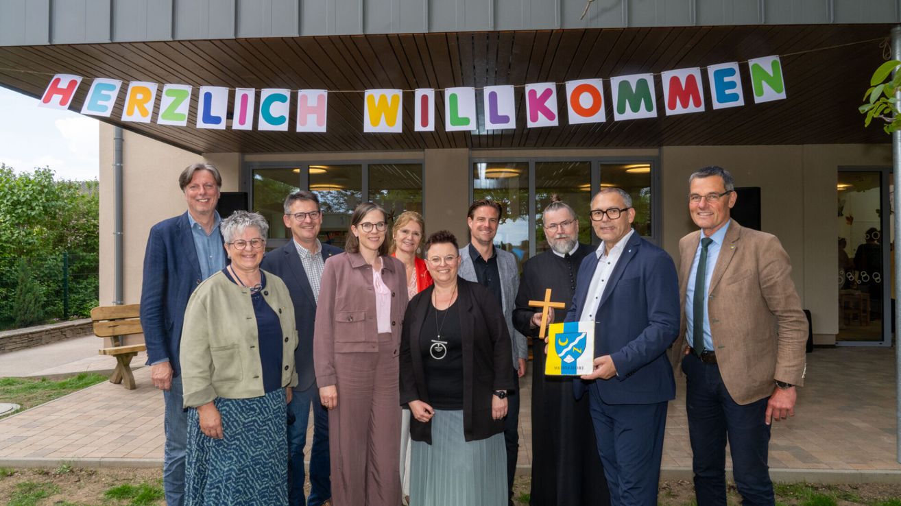 Eine Gruppe von Menschen steht vor einem Gebäude mit einem Banner, auf dem 'LICH WILLKOMM' steht. Sie lächeln und posieren für ein Foto.