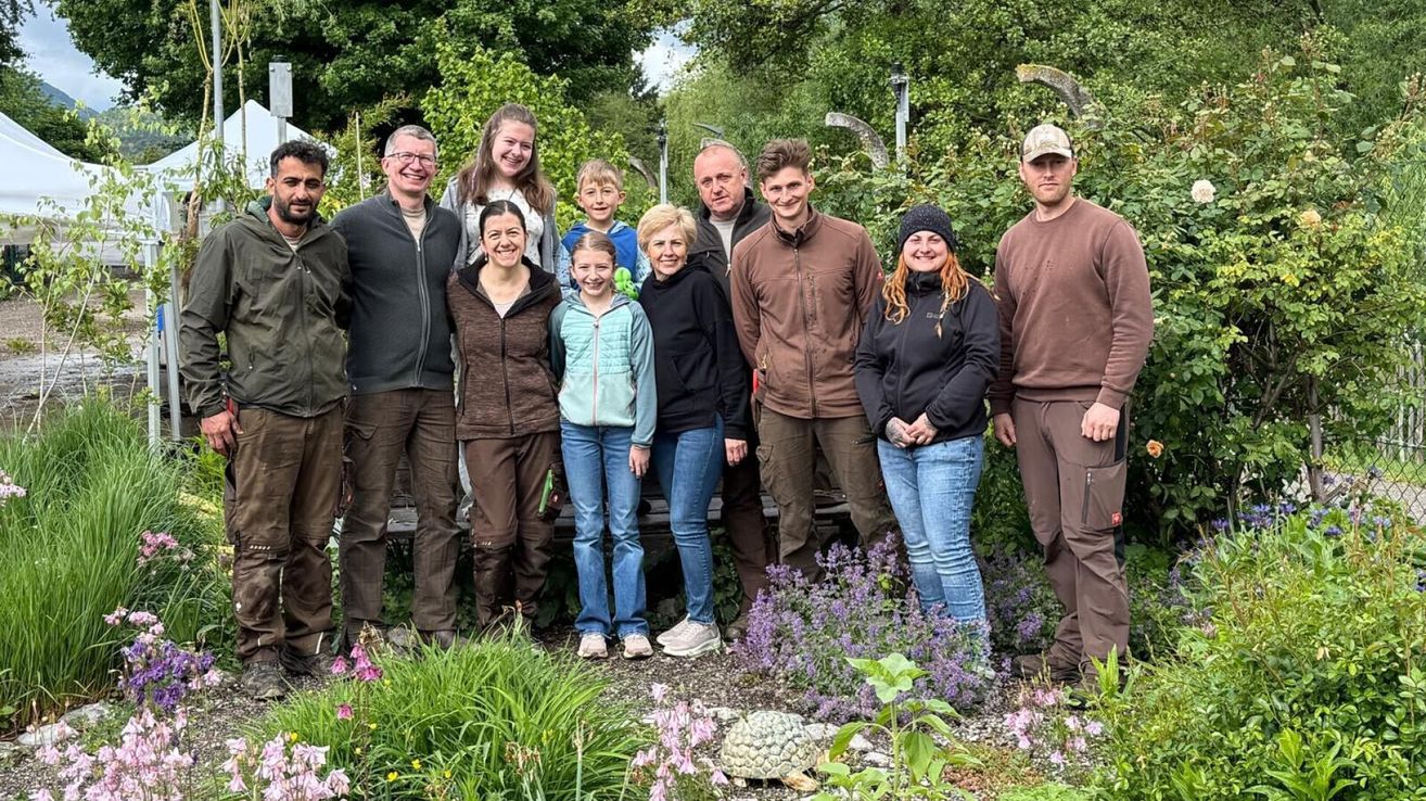 Eine neunköpfige Familie posiert für ein Foto im Garten. Sie stehen vor grünen Büschen und Blumen. Einige von ihnen tragen Hüte und Brillen.