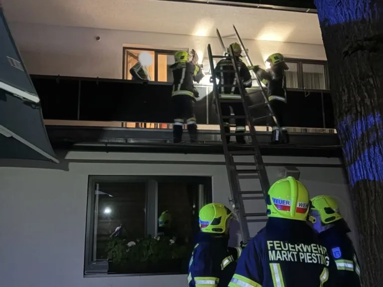 Eine Gruppe von Feuerwehrleuten befindet sich auf einem Balkon und versucht, jemanden aus einem Gebäude zu retten. Sie tragen Helme und Uniformen. Das Gebäude hat Glasfenster und eine Tür.