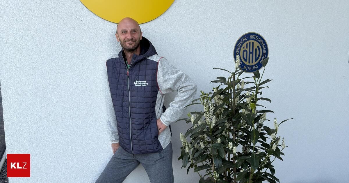 Ein lächelnder Mann mit einer blauen Weste steht an einer weißen Wand mit einem gelben Kreis und posiert neben einer grünen Pflanze mit weißen Blüten.