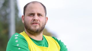 Ein Mann mit Bart und Schnurrbart in einem gelben und grünen Shirt schaut mit leicht geöffnetem Mund in die Kamera.