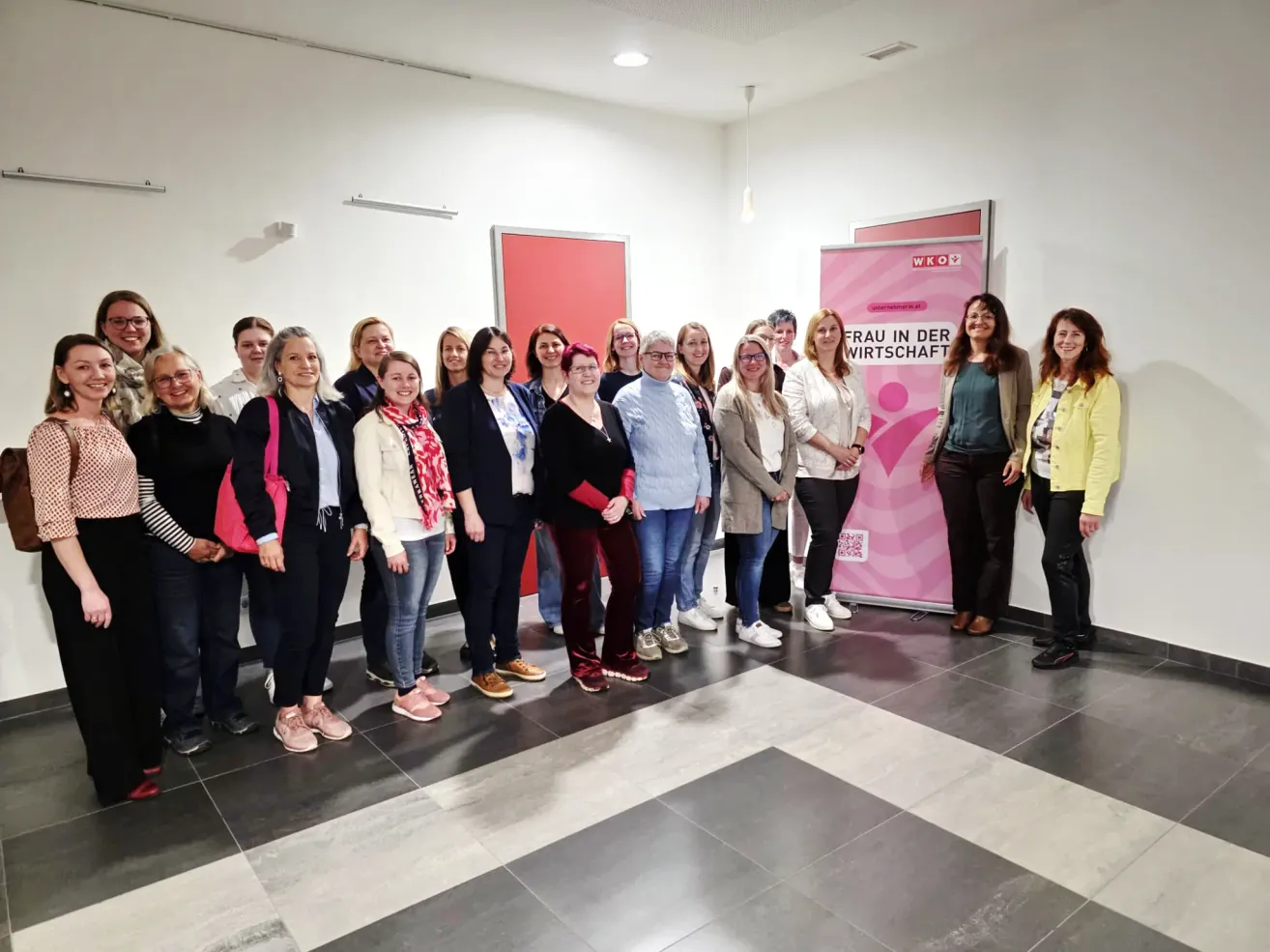 Eine Gruppe von Frauen in Geschäftskleidung posiert für ein Foto vor einem rosafarbenen Banner mit der Aufschrift 'Frau in der Wirtschaft'.