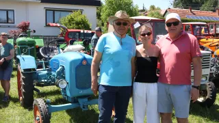 Drei Personen posieren für ein Foto vor einem blauen Traktor an einem sonnigen Tag. Hinter ihnen sind andere Fahrzeuge auf Gras geparkt. Ein Mann trägt einen Cowboyhut.