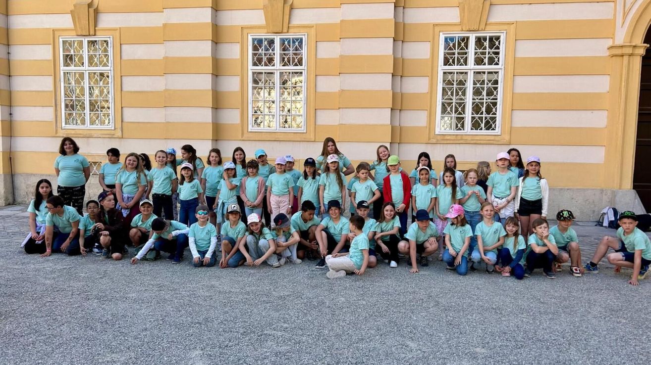 Eine Gruppe junger Menschen in passenden hellgrünen T-Shirts und Mützen posiert für ein Foto vor einem Gebäude mit gelben und weißen Streifen und großen Fenstern.