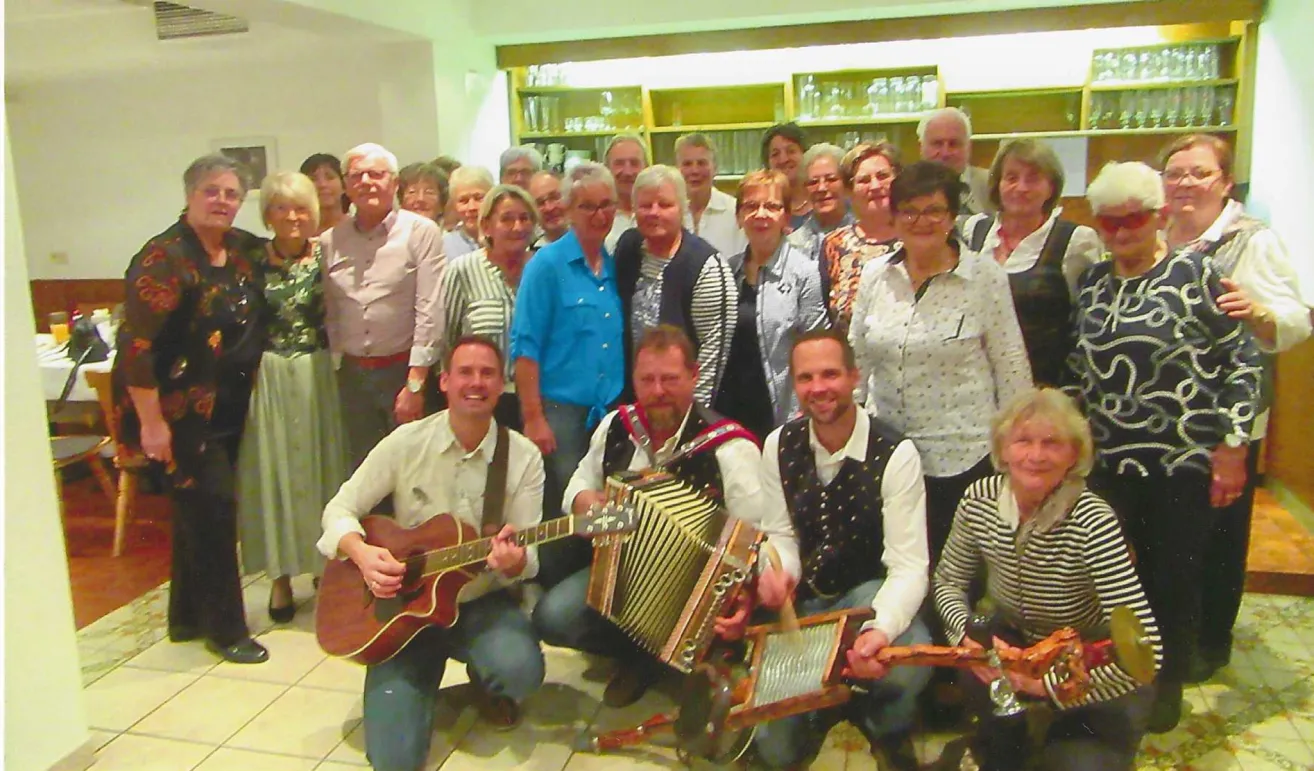 Eine Gruppe von Menschen in Freizeitkleidung posiert für ein Foto, einige mit Musikinstrumenten. Sie scheinen sich bei einer geselligen Zusammenkunft in einem gut beleuchteten Raum zu befinden.