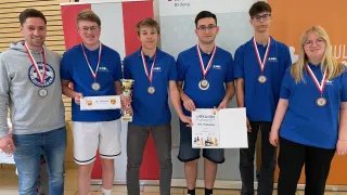 Fünf junge Männer in blauen Hemden mit Medaillen um den Hals stehen vor einem Banner. Sie halten einen Pokal und ein Zertifikat. Hinter ihnen ist eine Wand mit einem Logo.