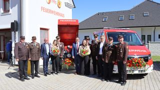 Eine Gruppe von Menschen, darunter Frauen mit Blumen, steht vor einem Feuerwehrhaus und einem Feuerwehrauto.