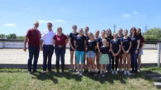 Eine Gruppe von Menschen steht zusammen auf dem Gras, wahrscheinlich für ein Foto. Zwei Männer und eine Frau tragen karmesinrote Shirts; der Rest, hauptsächlich Frauen, trägt Blau. Dahinter steht eine Metallbarriere.