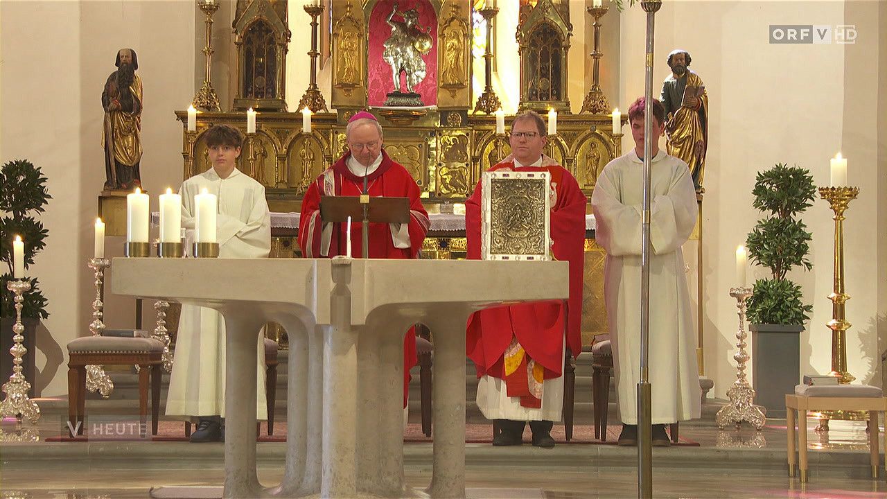 Eine religiöse Zeremonie in einer Kirche. Ein Priester liest aus einem Buch am Altar, begleitet von Messdienern und einem Jungen.