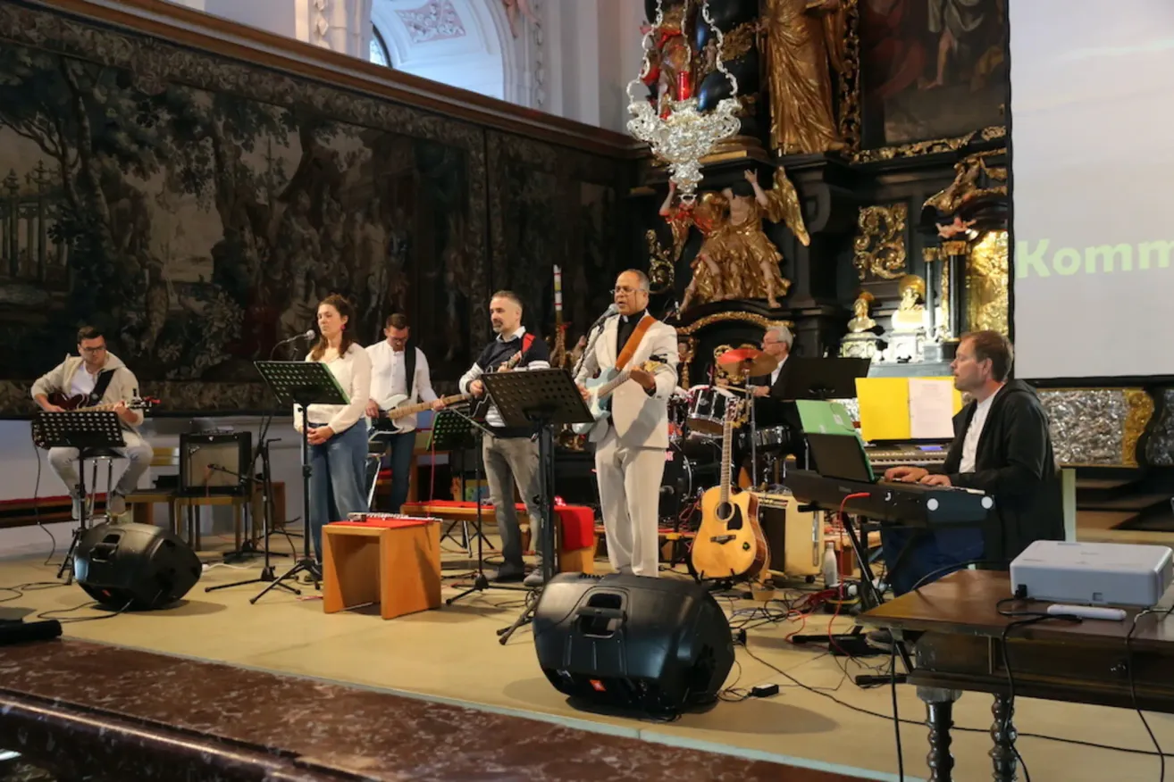 Eine Band tritt in einer Kirche auf. Sie spielen Gitarren und Trommeln, und einer singt. Dahinter befindet sich eine große Wand mit Gemälden.