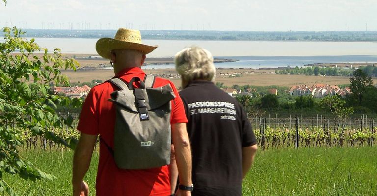 Zwei Männer stehen auf einem grasbewachsenen Hügel und blicken auf eine große, malerische Landschaft mit einem Gewässer, einem Weinberg und entfernten Häusern. Einer trägt ein rotes Shirt und einen Strohhut, der andere ein schwarzes Shirt mit weißem Text.