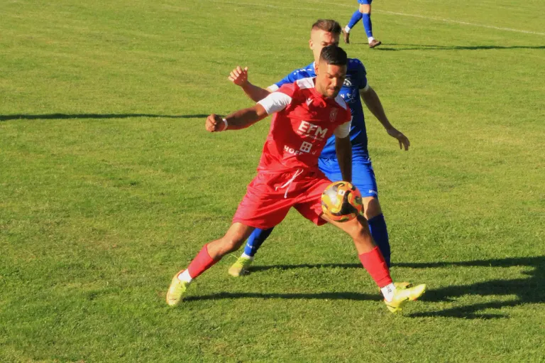 Ein Fußballspieler in Rot tritt den Ball, während ein anderer Spieler in Blau hinter ihm auf einem grünen Feld steht.