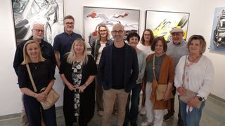 Eine Gruppe von Menschen steht vor Kunstgemälden, lächelt und posiert für ein Foto. Einige tragen Brillen und Jacken, und zwei Frauen tragen Halsketten. Die Gemälde sind groß und zeigen Autos.