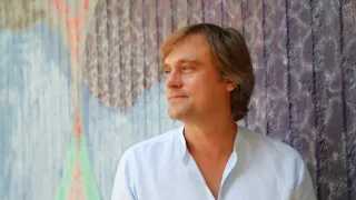 Ein Mann mit kurzen braunen Haaren in einem hellblauen Hemd vor einer farbenfrohen Wandmalerei.