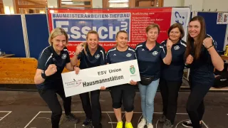 Eine Gruppe von Frauen steht zusammen und hält ein weißes Banner mit dem Text ESV Union Hausmannstatten. Sie lächeln und scheinen zu feiern. Die Umgebung ist drinnen, mit einem Banner im Hintergrund.