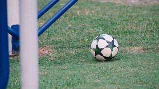 Ein Fußball mit grünem und schwarzem Design liegt auf einem Rasenfeld mit einem blauen Torpfosten auf der linken Seite.