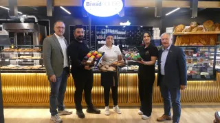Fünf Personen stehen vor einem Tresen mit der Aufschrift Bread House. Sie halten alle Tabletts mit Brot und Gebäck. Die beiden Männer in Anzügen lächeln.